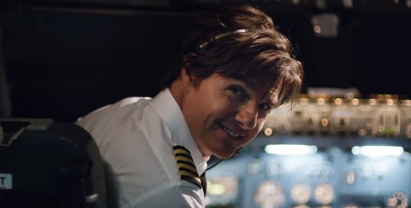 Tom Cruise in a the cockpit of a plane turning and smiling.